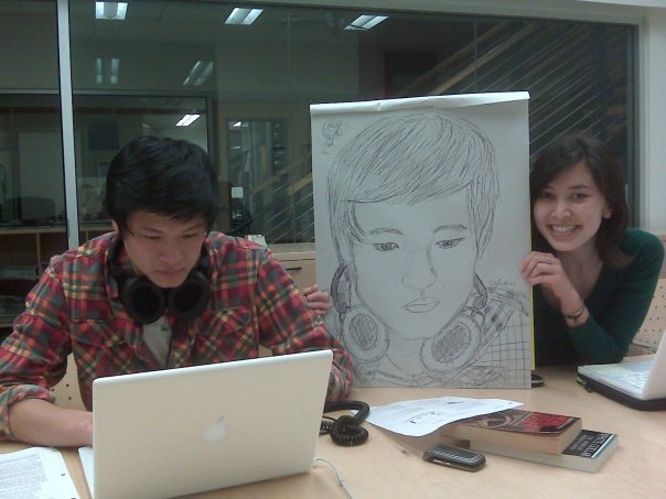 A man with headphones around his neck sits at his laptop. A woman beside him holds up a large sketch of the man in the same pose.